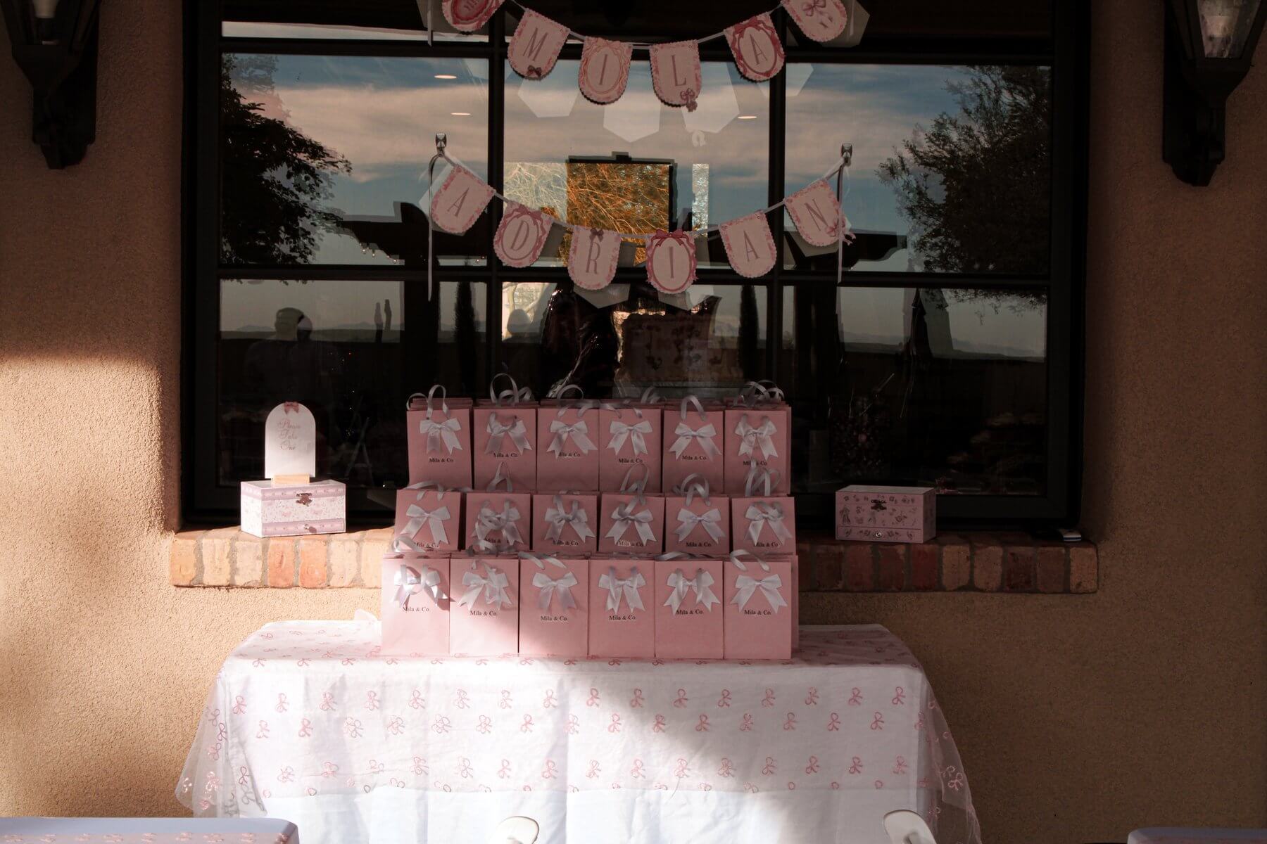 Pyramid of pink Mila & Co favor bags with white satin bows, styled on a bow-pattern tablecloth beneath custom hanging banners