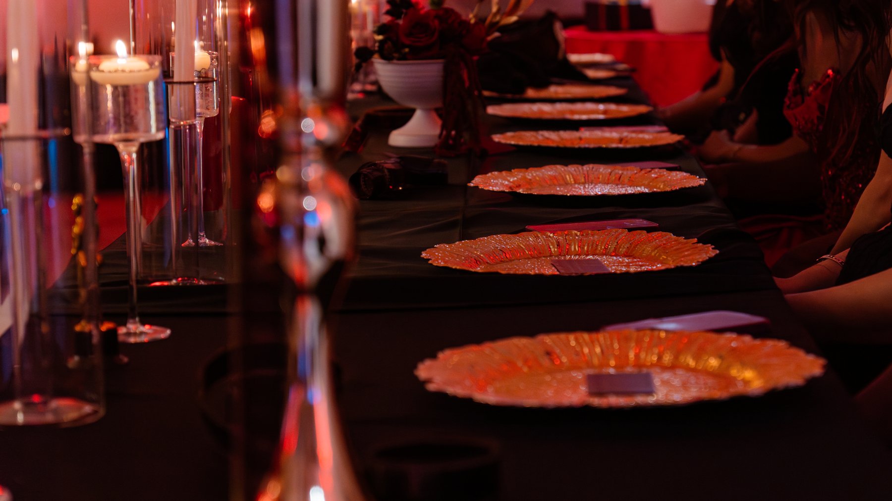 Long banquet table set with charger plates, candle vignettes, and red and black Hollywood gala styling under dramatic low light