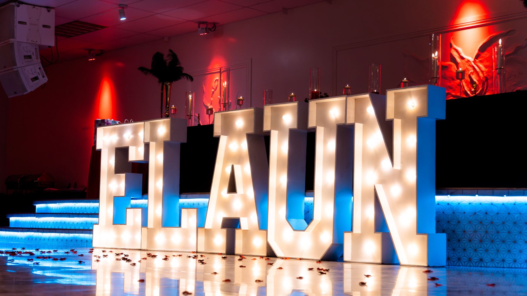 Giant illuminated ELAUN marquee letters set in front of the head table at a Hollywood-themed graduation gala, with feather centerpieces and dramatic uplighting