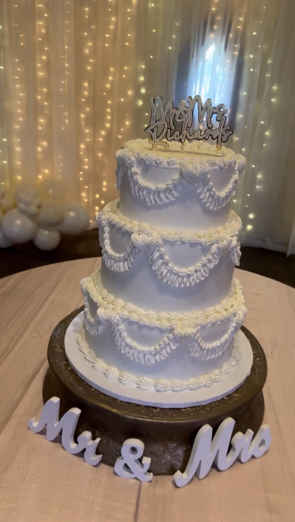 Three-tier white Mr and Mrs Richards wedding cake with cascading white florals and gold topper